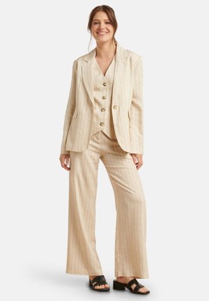 Beige gestreifter Anzug mit einem strukturierten Blazer, einer geknöpften Weste und weitem Hosenbein. Das Material wirkt leicht und texturiert. Schwarze Sandalen.