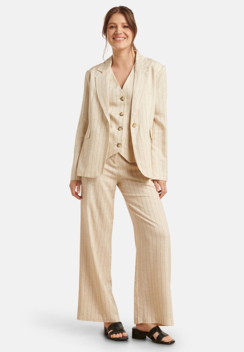 Beige streepjasje met een gestructureerde blazer, knopenvest en wijde broekspijpen. Materiaal lijkt licht en textuur te hebben. Zwarte sandalen.