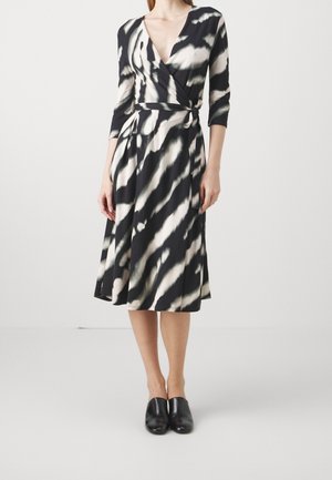 Kobieta w czarno-białej sukience o wzorze zebry, z rękawami do połowy przedramienia, stojąca na tle białego tła, w czarnych butach wsuwanych.