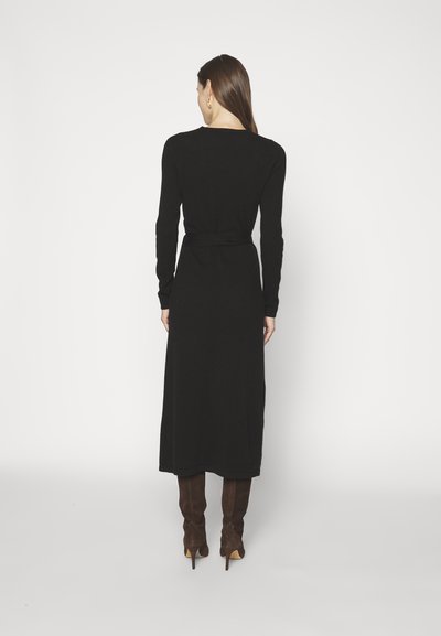 Robe noire en maille avec des manches longues, une taille cintrée et une jupe fluide arrivant au milieu du mollet. Portée avec des bottes en cuir suédé marron jusqu'aux cuisses.