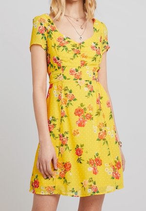 Femme portant une robe jaune à manches courtes avec un imprimé floral rose et orange et des colliers superposés, debout devant un fond gris uni.
