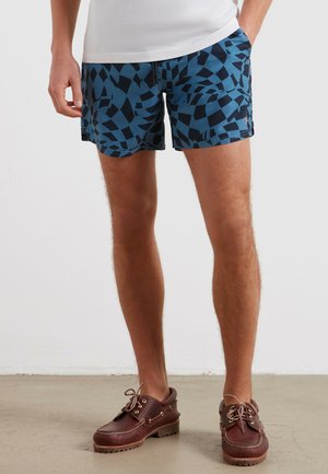 Mężczyzna stojący z rękami w kieszeniach, ubrany w niebieskie kąpielówki w wzory i brązowe skórzane buty żeglarskie, stoi na betonowej podłodze.