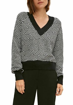 Femme portant un pull à col en V à motif géométrique noir et blanc avec poignets et ourlet noirs, associé à un pantalon noir.