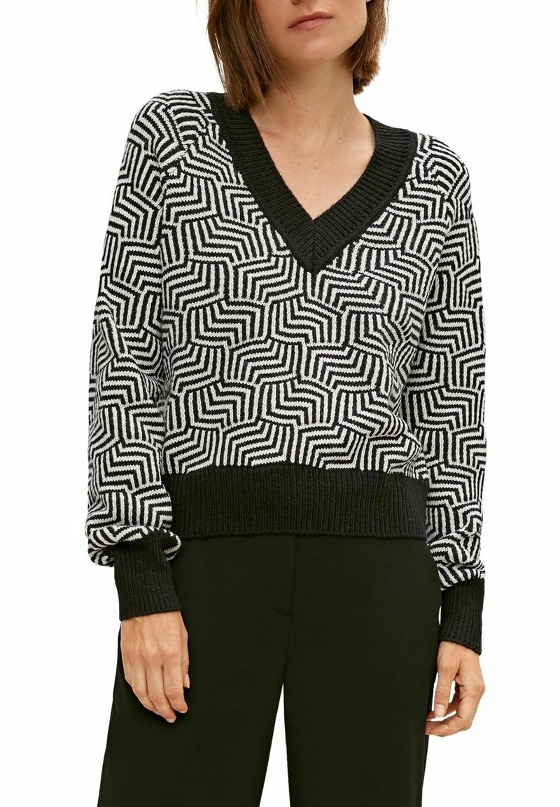 Femme portant un pull à col en V à motif géométrique noir et blanc avec poignets et ourlet noirs, associé à un pantalon noir.