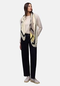 Femme portant un pull beige, un jean large foncé, des baskets blanches, et une grande écharpe ivoire avec des motifs noirs et jaunes drapée sur les épaules.