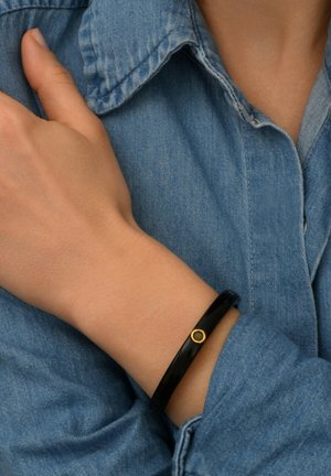 Bracelet noir en matériau lisse, avec un accent circulaire en or. Porté au poignet contre une chemise en denim bleu.