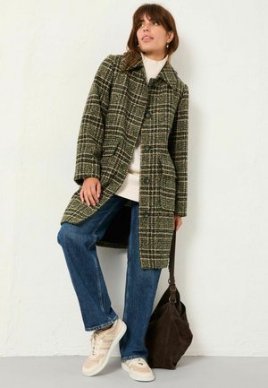 Plaid coat in dark green and beige with a collar, button front, and two pockets, worn over a cream knit sweater and blue jeans. Accessorized with a dark bag.