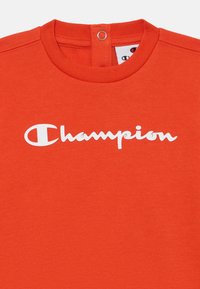 Sarkans kokvilnas T-krekls ar apaļu izgriezumu, aizmugurē ar pogu detaļu un baltu "Champion" logo uz krūškurvja.