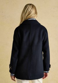 Tumedast merest villane koot, millel on väljendunud disain, suur krae ja nööpidega mansetid, sile tekstuur ja sirge lõige.