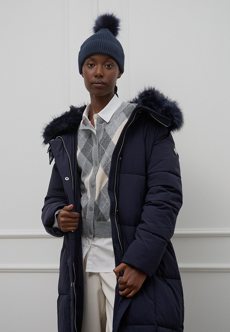 Cappotto imbottito blu navy con cappuccio bordato in pelliccia, abbinato a un maglione grigio a rombi e un berretto blu navy con pom-pom. Sfondo semplice.