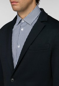 Muž má na sobě černý blejzr přes bílou košili s úzkými černými vertikálními pruhy, zapnutou až ke krku.