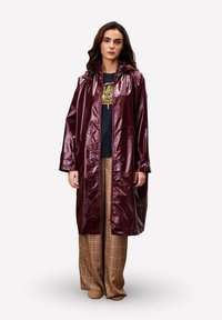 Bordeaux, glanzende regenjas met een kap en zakken; gedragen over een zwart graphic T-shirt en geruite wijde broek. Zachte textuur en oversized pasvorm.