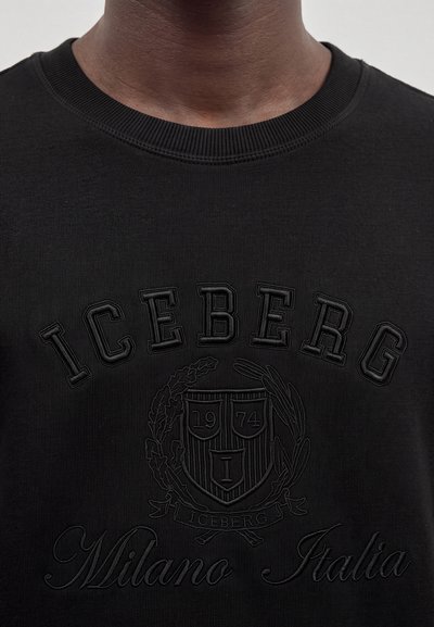 Крупен план на черен суитшърт с релефен текст "ICEBERG" и "Milano Italia" около детайлен гербов дизайн.