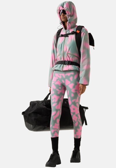 Person iført pink og grå camouflage hættetrøje og leggings, sorte støvler og solbriller, bærende en sort sportstaske og en sort rygsæk.
