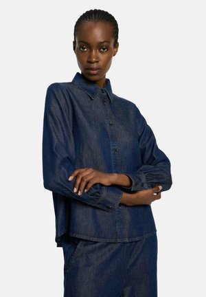 Woman wearing dark blue denim shirt and matching pants, standing with arms crossed against a plain white background.