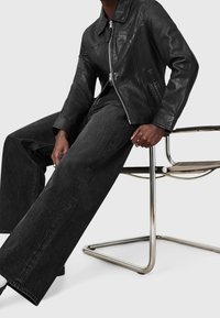 Veste en cuir noire à fermeture éclair frontale, col classique et poches latérales, associée à un jean en denim sombre à jambes larges et une chaise minimaliste.