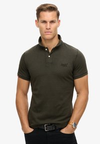Mørkegrøn polo t-shirt med ribbet tekstur, tre-knaps placket og et diskret logo på brystet. Ærmeafslutningerne har orange accenter.