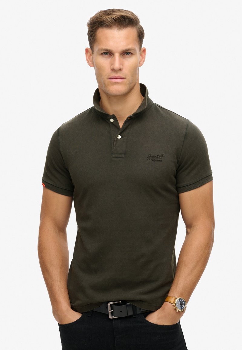Mørkegrøn polo t-shirt med ribbet tekstur, tre-knaps placket og et diskret logo på brystet. Ærmeafslutningerne har orange accenter.