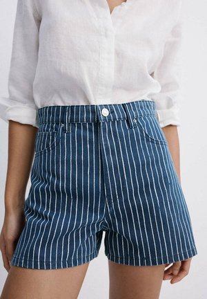 Personne portant un short en jean bleu avec des rayures blanches verticales et une chemise blanche à manches longues avec les manches retroussées.