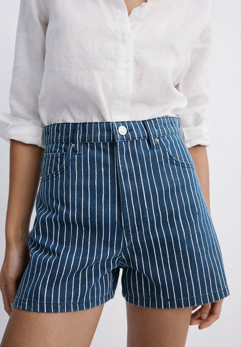 Personne portant un short en jean bleu avec des rayures blanches verticales et une chemise blanche à manches longues avec les manches retroussées.