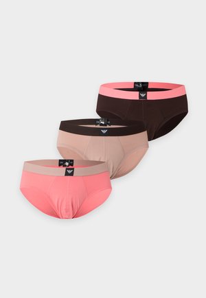 Homme aux cheveux foncés portant un slip rose clair avec une ceinture noire, debout pieds nus sur un fond blanc uni.