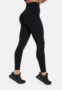 Hoge taille zwarte leggings gemaakt van rekbaar materiaal, met een naadloos ontwerp en een logo-accent op het onderbeen. Gecombineerd met zwarte sneakers.