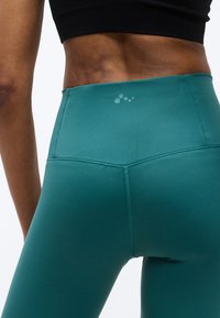 Κοντινή λήψη από το κάτω μέρος της πλάτης και τη μέση ενός ατόμου που φοράει πετρόλ κολάν ψηλόμεσο και μαύρο κοντό τοπ, με λευκό φόντο.