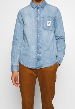 Mężczyzna w jasnoniebieskiej koszuli jeansowej zapinanej na guziki z małą naszywką z logo na kieszeni piersiowej i brązowych spodniach, stojący z jedną ręką w kieszeni.