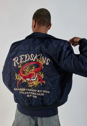 Homme portant une veste bleu marine avec un dragon rouge brodé et le texte "REDSKINS DRAGON TEDDY BY RDS DA-NANG HUE 67-70" au dos.