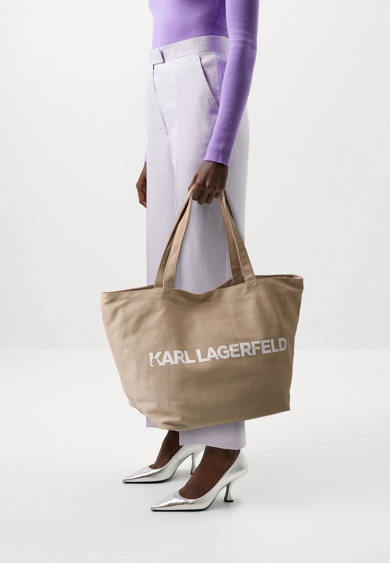 Kaki tygväska med "KARL LAGERFELD"-logotyp i vitt. Tillverkad av strukturerad textil. Modellen bär lavendelfärgade byxor och silverfärgade högklackade skor.