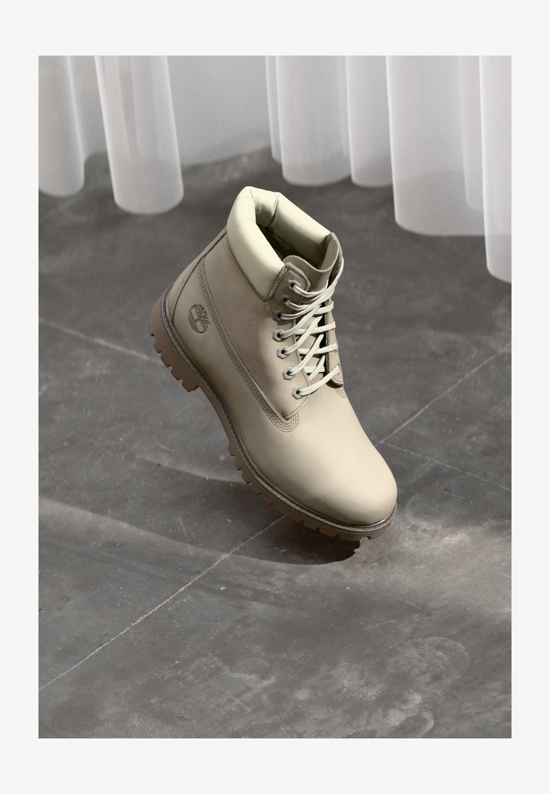 Botín de trabajo beige con cordones, con cuello acolchado y suela gruesa, sobre un suelo de baldosas grises bajo cortinas blancas de gasa.