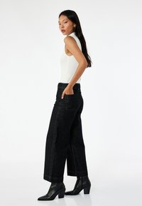 Zwarte denim wijde pijpen broek met voorzakken, gecombineerd met een strakke witte mouwloze top. Model poseert zijwaarts tegen een neutrale achtergrond.