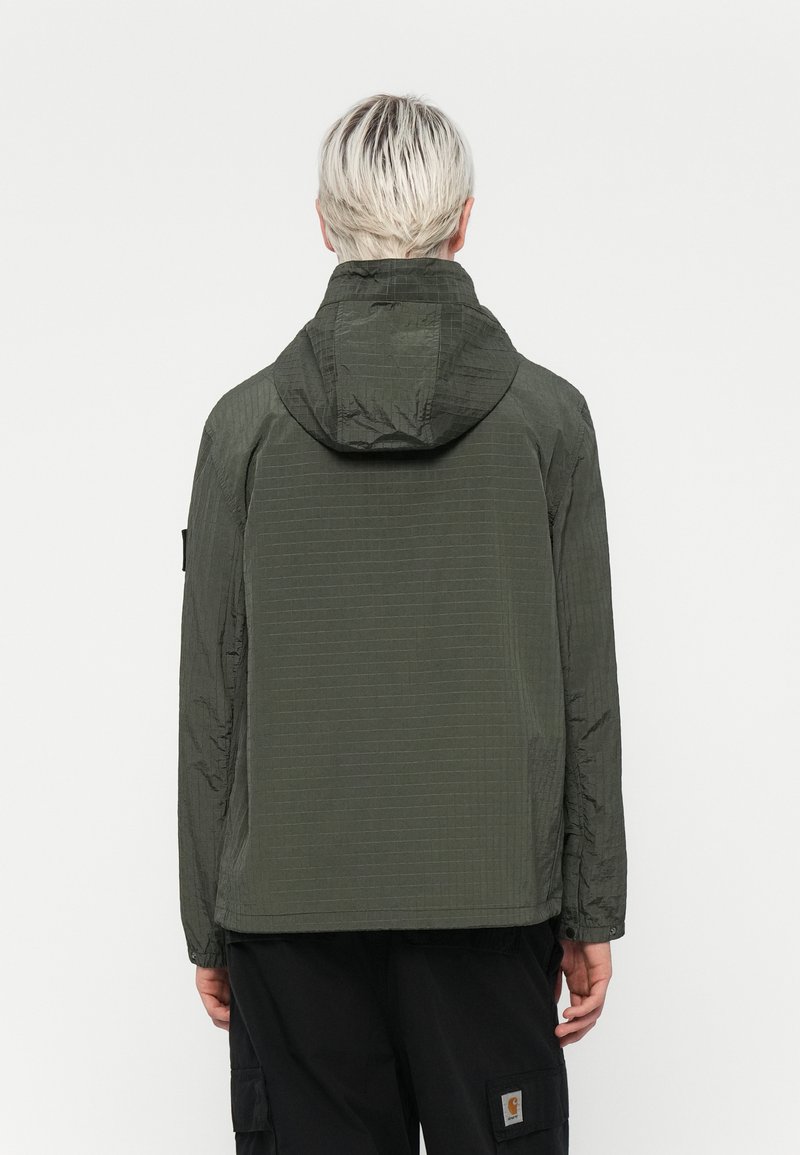 Jachetă ușoară de culoare verde închis, cu glugă. Țesătură texturată cu aspect crăpat, cusături vizibile și manșete elastice. Design simplu și funcțional.