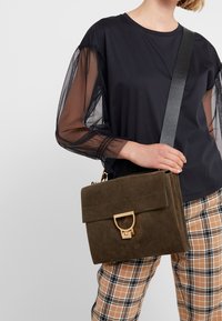 Zielona zamszowa torebka na ramię z złotym zapięciem, o prostokątnym kształcie, krótką klapką i teksturowanym paskiem na ramię, noszona z czarnym topem i kraciastymi spodniami.