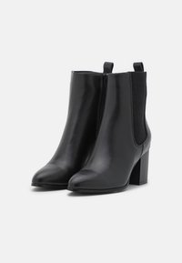 Par de botas de piel negra por el tobillo con tacones de bloque, cremallera lateral y paneles elásticos sobre un fondo blanco.