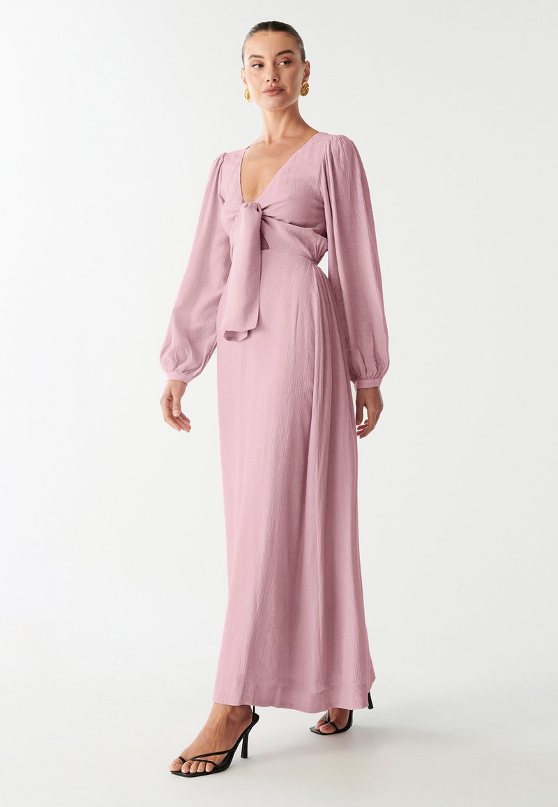 Woman wearing a long, light pink dress with puffed sleeves, deep V-neck, and a front tie, paired with black high-heeled sandals.