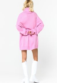 Převeliký růžový svetrový šaty s vysokým límcem, žebrovanými manžetami a volným střihem. Nošeny s bílými kozačkami sahajícími ke kolenům.