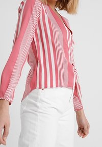 Rosa- und weiß gestreifte Bluse mit langen Ärmeln und V-Ausschnitt, aus leichtem Stoff gefertigt; mit einem verdrehten Frontdetail.