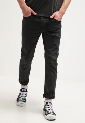 Homme portant un pantalon noir retroussé, des baskets montantes noires et blanches, et une chemise noire, les mains dans les poches, debout sur un sol blanc.