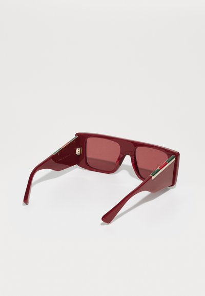 Ochelari de soare roșii cu rame dreptunghiulare, lentile tintate și finisaj lucios. Caracteristici hardware de tonalitate aurie și un accent tricolor pe brate.