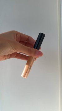 Correttore cosmetico in un tubo di vetro trasparente con una tonalità pesca-beige, dotato di un tappo a vite nero e un applicatore a forma di piede di cerbiatto.