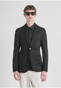 Blazer in suede nero con risvolto, chiusura a un bottone, due tasche frontali e una trama sottile, abbinato a una camicia nera e occhiali da sole.