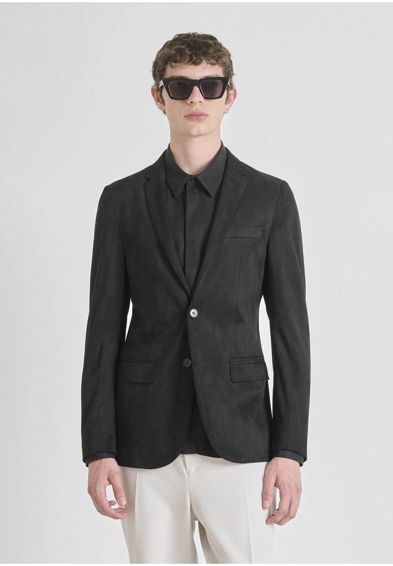 Blazer in suede nero con risvolto, chiusura a un bottone, due tasche frontali e una trama sottile, abbinato a una camicia nera e occhiali da sole.