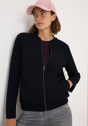 Schwarze Zip-Jacke mit Stehkragen, dehnbarem Stoff und elastischem Bund. Kombiniert mit einem dunkelvioletten Oberteil und grauen Jeans. Helle rosa Mütze.