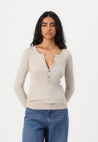 Bluză bej cu mânecă lungă, cu textură cu nervuri, având un guler decupat și trei butoane la gât; purtată cu blugi din denim albastru.