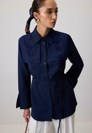 Camisa azul marino con detalles florales bordados en los hombros, con botones en la parte delantera y cinturonado en la cintura. Mangas largas con puños acampanados.