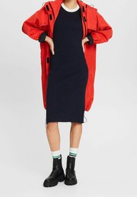 Veste imperméable rouge par-dessus une robe côtelée bleu marine, associée à des bottes noires épaisses et des chaussettes blanches avec des rayures vertes.
