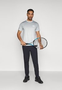 Heren sportoutfit met een kortärmige wit- en grijsverloopshirt met stippenpatroon, gecombineerd met zwarte sportbroeken en zwarte sneakers.