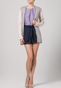 Lekki, otwarty kardigan w szaro-kremowym splocie, połączony z wzorzystą purpurowo-białą bluzką i granatowymi szortami z wysokim stanem.