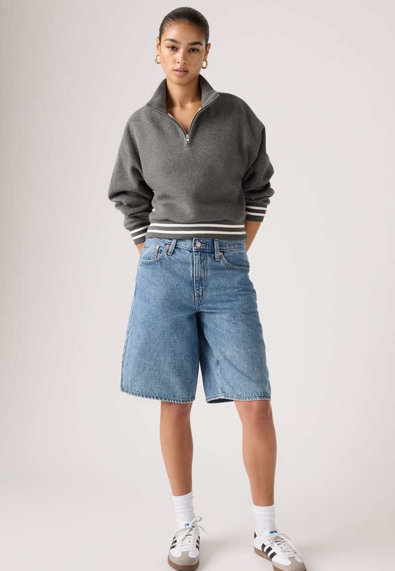 Jeune femme portant un sweat-shirt gris à fermeture éclair, un long short en denim, des chaussettes blanches et des baskets blanches, debout devant un fond uni.